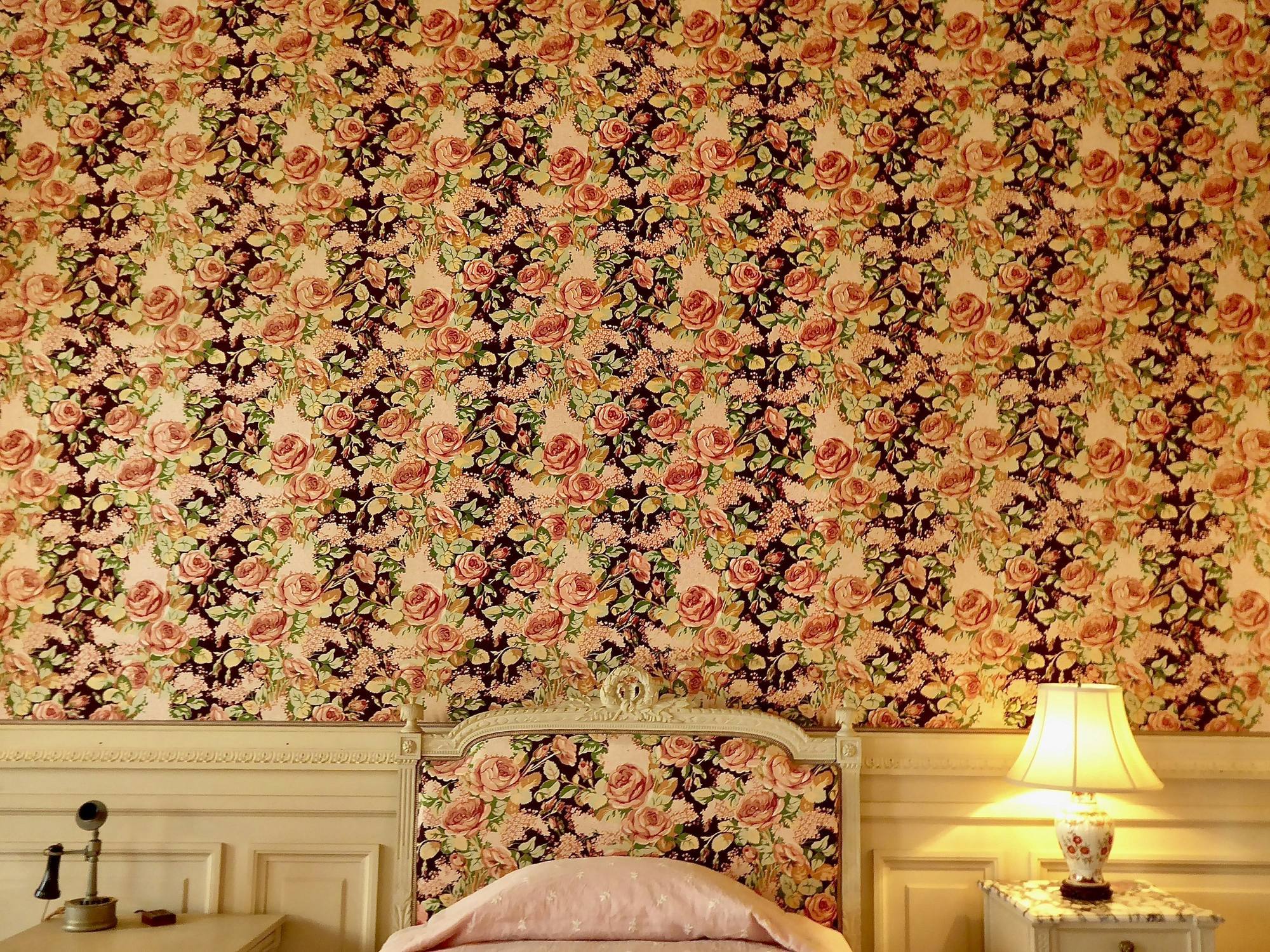 Residential bedroom feature wall covered in richly patterned rose wallpaper, with a matching upholstered headboard.